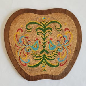 May include: Apple-shaped coaster with a decorative design. The coaster features a brown wooden frame and a cork center. The center has a colorful floral and bird design in green, blue, yellow, and red.