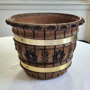 May include: A brown wooden planter with a carved floral design and two brass bands. The planter has a barrel-like shape and is suitable for indoor or outdoor use.