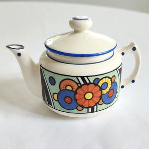 May include: A small, cream-colored ceramic teapot with a lid and handle. The teapot is decorated with a colorful floral design in orange, blue, and yellow, set against a green and black background. Blue accents are on the rim and handle.