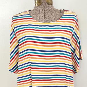 May include: A colorful, striped tunic dress displayed on a mannequin. The dress features horizontal stripes in red, yellow, orange, blue, and teal, alternating with white stripes. The dress has short sleeves and a rounded neckline, offering a vibrant and casual look.