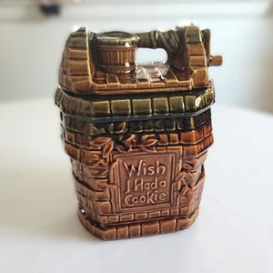 May include: A brown ceramic cookie jar shaped like a wishing well with a brick-textured exterior. The front features a raised rectangular panel with the embossed text "Wish I Had a Cookie." The lid includes a small bucket and a crank handle detail.