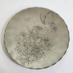 May include: A round, gray decorative stone featuring an embossed design of a cluster of daisies and two butterflies. The stone has a textured surface and a scalloped edge, suitable for garden decor.