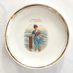 May include: A vintage white ceramic plate with a gold rim. The plate features a colorful illustration of a woman in a blue dress standing near the ocean. The text "DOWNLINGS STS OF M. CAMPBELL" is printed on the plate.
