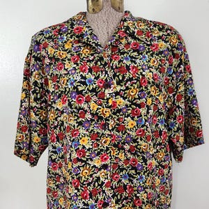 May include: A short-sleeved shirt with a vibrant floral pattern. The shirt features a black background covered in a colorful array of red, yellow, purple, and blue flowers. The shirt is displayed on a mannequin.
