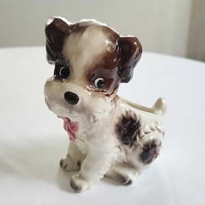May include: A ceramic puppy figurine with brown and white markings, a pink bow, and a small opening on its back. The puppy is sitting on its hind legs, with its front paws extended. The figurine has a glossy finish.