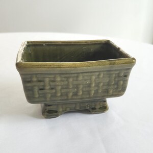 May include: A small, square, ceramic planter with a woven basket design in a muted green color. The planter sits on a pedestal base with three small feet. The interior of the planter is a darker shade of green.
