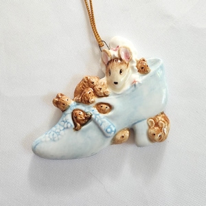 May include: A ceramic ornament featuring a light blue shoe filled with several brown mice. The shoe has a decorative bow and is accented with gold trim. A tag with text hangs from a gold string.