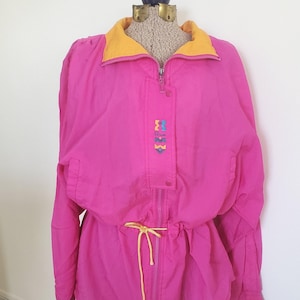 May include: A bright pink vintage jacket with a yellow collar and drawstring waist. The jacket has a zipper closure and a small decorative emblem with colorful shapes. The jacket is displayed on a mannequin.