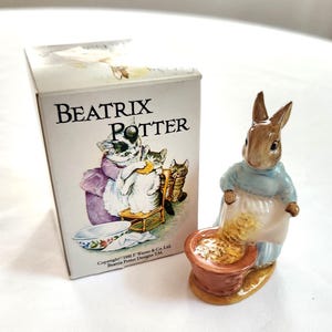 May include: A Beatrix Potter figurine of a brown rabbit wearing a blue shawl and apron, pouring from a brown bowl. The figurine is next to a box with the words "Beatrix Potter" and an illustration of cats.