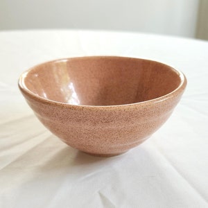 May include: A small, light pink ceramic bowl with a speckled texture. The bowl is empty and sitting on a white tablecloth.
