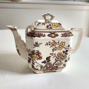 May include: A white teapot with a square body and a floral pattern in shades of brown, yellow, and green. The teapot has a lid with a knob on top.