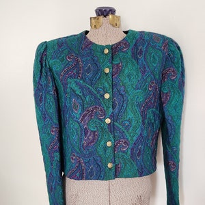 May include: A teal, blue, and purple paisley patterned jacket with gold buttons. The jacket has long sleeves and is displayed on a mannequin. The jacket's design features a repeating teardrop-shaped motif.