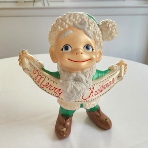 May include: A ceramic Christmas elf figurine wearing a green and white outfit and a white fur hat. The elf is holding a scroll that says "Merry Christmas".