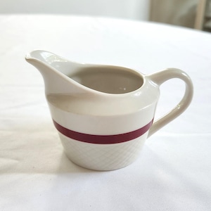 May include: A white ceramic creamer with a burgundy stripe around the middle. The creamer has a rounded body and a small spout.