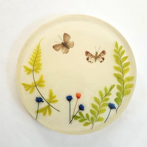 May include: A round, cream-colored coaster with a botanical design. Two butterflies and sprigs of green ferns and blue and orange flowers are embedded in the resin. The coaster is a decorative item.