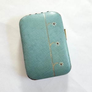 May include: A vintage, rectangular, teal-colored case with a textured surface. The case features a gold floral design with three small white flowers. The case has a gold clasp on the side and is likely used for carrying small items.