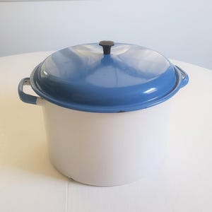 May include: A white enamel pot with a blue lid and handles. The lid has a black knob. The pot is sitting on a white surface. The pot is likely used for cooking.