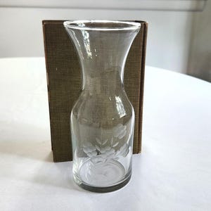 May include: A clear glass water carafe with a flared top and etched floral design. The carafe is positioned in front of a brown rectangular object, likely a book or decorative piece. The carafe sits on a white surface.