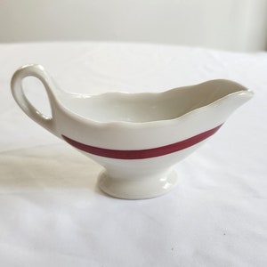 May include: A white ceramic gravy boat with a single red band around the middle.