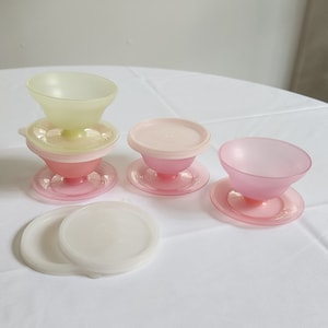 May include: A collection of vintage plastic dessert dishes in pastel colors. The set includes pink, light pink, and yellow-green bowls with matching saucers and lids. The bowls are stacked and arranged on a white surface.