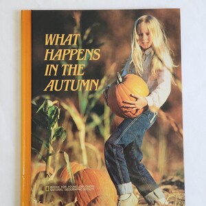 May include: A book cover with the title "WHAT HAPPENS IN THE AUTUMN" in gold lettering. The cover features a young person holding a large orange pumpkin. The book is part of the "BOOKS FOR YOUNG EXPLORERS" series.