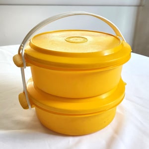 May include: A yellow two-tiered lunch container with a white handle. The container is made of plastic and has a round shape.