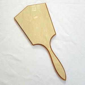 May include: A hand mirror with a light beige, geometric-shaped head and a long, slender handle. The mirror has a warm, neutral color palette and a smooth, polished finish. The handle and the border of the mirror are a darker shade of beige.