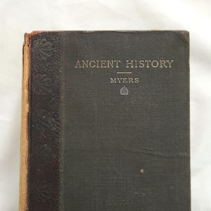 May include: A vintage book titled "ANCIENT HISTORY" by Myers. The book has a dark green cover with gold lettering and a decorative brown spine. The edges of the pages are aged, giving it a classic look.