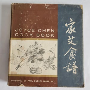 May include: A vintage cookbook titled "Joyce Chen Cook Book" with a brown and blue cover. The cover features the title in white text, along with an illustration of vegetables and Chinese characters. The book is slightly worn.
