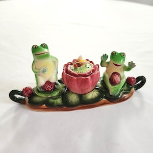 May include: A ceramic frog-themed centerpiece featuring two green frogs flanking a pink lotus flower with a small dish inside. The frogs and flower sit on a green leaf-shaped base with red accents. The centerpiece is on a white surface.