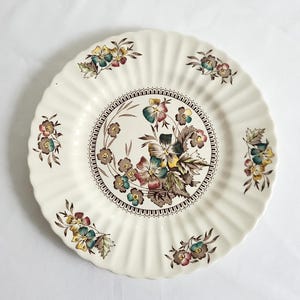May include: A round, cream-colored ceramic plate with a scalloped, fluted edge. The center features a detailed floral bouquet in shades of brown, blue, and yellow, surrounded by a decorative border. Smaller floral clusters repeat around the rim.
