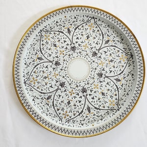 May include: Round decorative tray with a floral pattern in white, gray, and gold. The tray has a gold rim with a geometric design. The center of the tray features a white circle. The tray is on a white background.