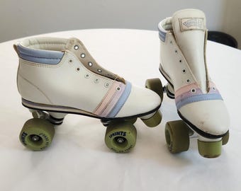 May include: Pair of vintage roller skates. The skates are white with light blue and pink stripes and green wheels. The skates are sitting on a white surface.