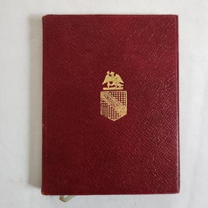 May include: A small, rectangular, burgundy-colored book with a gold emblem on the front. The emblem features a shield and a bird. A silver bookmark is visible at the bottom edge of the book.