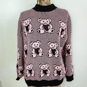 May include: Pink and black striped sweater featuring repeating teddy bear designs. The sweater has a black turtleneck, cuffs, and hem. The teddy bears are pink with black accents.