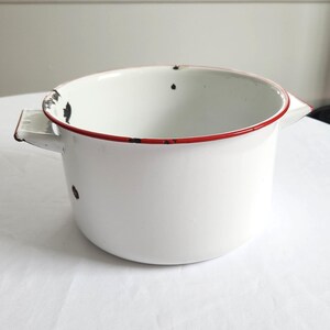May include: White enamelware pot with a red rim and two side handles. The pot has some black spots and wear, giving it a vintage look. A classic kitchenware item.