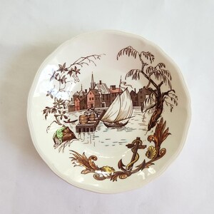 May include: A round, cream-colored ceramic dish with a scalloped edge. The center features a detailed brown and green illustration of a harbor scene with a sailboat, buildings, and barrels, accented by a decorative anchor and foliage at the bottom.