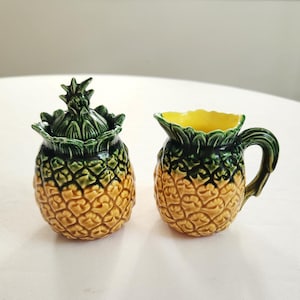 May include: A ceramic pineapple-shaped sugar bowl with a lid and a matching creamer. The sugar bowl has a green leafy top and a yellow and brown body. The creamer has a green handle and a yellow interior.