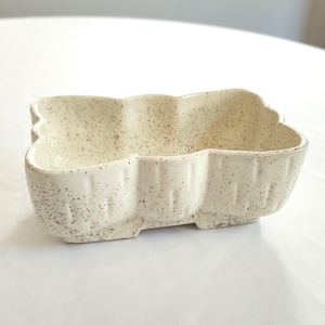 May include: A speckled, off-white ceramic dish with a unique, scalloped shape. The dish is divided into three sections and has a textured surface. This decorative piece is suitable for serving snacks or as a small decorative item.