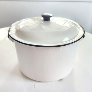 May include: A white enamel pot with a lid. The pot has a black rim and handle, and a black knob on the lid. The pot is sitting on a white surface. The pot is a classic kitchenware item.