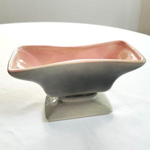 May include: A ceramic dish with a square base and a flared, rectangular bowl. The bowl is pink inside and transitions to gray on the outside. The base is a light gray color. The dish is likely for serving food or displaying decorative items.