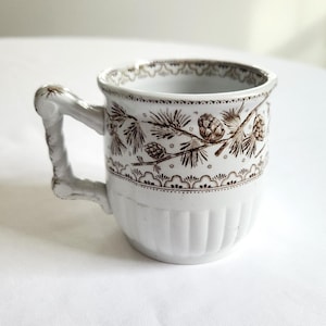 May include: A white ceramic mug featuring a brown transferware pattern of pine cones and needles around the upper rim. The mug has a textured, fluted base and a uniquely shaped handle that resembles a twisted branch or twig design.