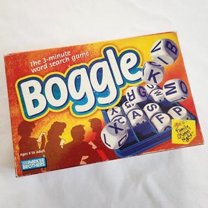 May include: Boggle word search game box with orange and yellow background. The box features the game's name in large blue letters and includes dice with letters. The box also includes the text "The 3-minute word search game."