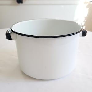 May include: A white enamel pot with a smooth finish and a black rim. The cylindrical cooking vessel features two small black side handles attached to the upper exterior. The interior of the pot is white, and the overall design is simple and functional.