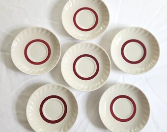 May include: Six white ceramic saucers with a red ring around the center. The saucers are arranged in a circle on a white tablecloth.