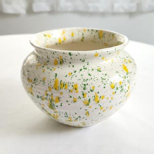 May include: A small, round ceramic bowl with a wide opening and a flared rim. The exterior features a white base color decorated with a speckled pattern of vibrant yellow and forest green paint splatters distributed across the entire surface.