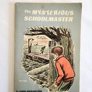 May include: Vintage book cover titled "The Mysterious Schoolmaster" with a light blue background. The cover features an illustration of a boy in an orange jacket, looking at a pier scene. The author's name, Karin Anckarsvärd, is also visible.