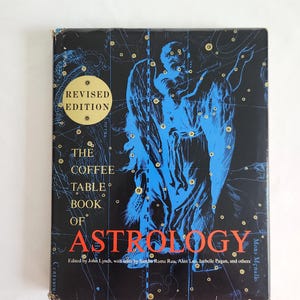 May include: A hardcover book titled The Coffee Table Book of Astrology, edited by John Lynch. The black cover features a blue celestial illustration of a winged figure and star charts, with a gold circular sticker reading Revised Edition.