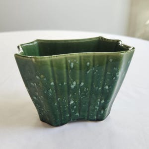 May include: A square, green ceramic planter with a textured, ridged design. The planter has a scalloped rim and a speckled finish, adding visual interest. This decorative piece is suitable for small plants or succulents.