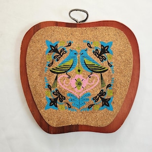 May include: Apple-shaped wall hanging with a cork center and a wooden frame. The cork features a colorful design of two blue birds facing each other, surrounded by floral accents in pink, blue, and black. A metal loop is attached for hanging.